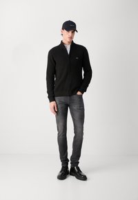 Maglione nero a costine con zip e un piccolo logo, abbinato a jeans slim-fit grigio scuro e scarpe nere stringate. Il modello indossa un cappellino nero.