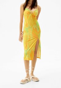 Maxi šaty v žluté barvě s potiskem palmových listů v zelené a oranžové barvě, s předním zavazováním a bočním rozparkem. Vyrobeno z lehkého a prodyšného materiálu.