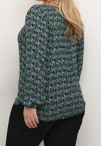 Haut à motif floral présentant des couleurs bleu-vert, blanc et noir, à manches longues et à coupe décontractée. Tissu texturé avec un détail froncé au dos.