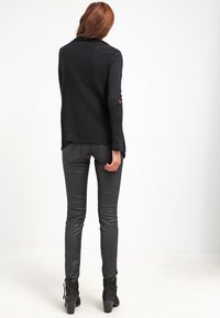 Cardigan noir en tricot avec un col, des manches longues ornées de boutons rouges, et une coupe décontractée, associé à un legging en cuir noir et des bottines.