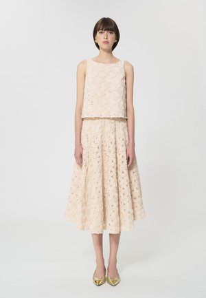 Donna in piedi con top smanicato color crema a trama e gonna midi abbinata con motivo floreale, indossa décolleté dorate con punta a punta, sfondo neutro.
