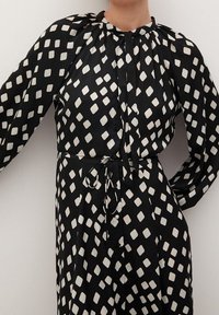 Vestido negro de manga larga con un patrón de rombos blancos, que presenta un escote fruncido y una cintura con cordón para un ajuste ajustable.