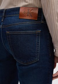 Traperice tamnoplave boje s glatkom teksturom. Sadrže vidljivu kožnu aplikaciju s "Calvin Klein" logotipom i kontrastnim šavovima.