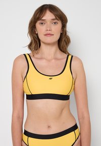 Żółty stanik sportowy z czarnymi wykończeniami i logo, z dekoltem w serek oraz szerokimi ramiączkami, wykonany z gładkiego, elastycznego materiału.