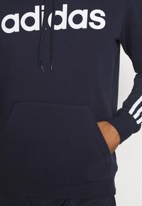 Haut en coton bleu marine avec le logo "adidas" blanc, poche kangourou à l'avant, capuche avec cordon de serrage et trois stripes blanches sur chaque manche.