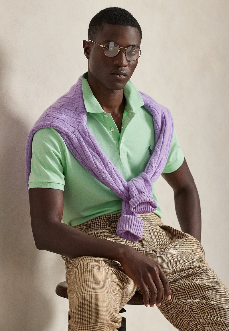 Ανοιχτό πράσινο πόλο πουκάμισο με  λιλά πλεκτό πουλόβερ δεμένο γύρω από τους ώμους, συνδυασμένο με μπεζ καρό παντελόνια. Λείες υφές και άνετη εφαρμογή.