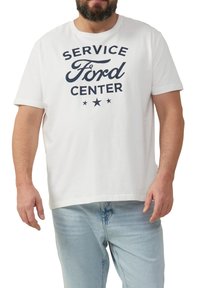 Man in een wit T-shirt met de tekst "Service Ford Center" en drie sterren, gecombineerd met lichtblauwe jeans, staande tegen een witte achtergrond.