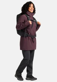 Bordeauxfarbene Outdoor-Jacke mit hohem Kragen und Reißverschluss, getragen über schwarzer Hose und einem schwarzen Rucksack, der verstellbare Riemen und Schnallen hat.