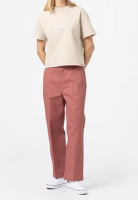 Béžové bavlněné zkrácené tričko s logem "Dickies", spárované s vysoko pasovými, rovnými kalhotami v růžové barvě a bílými teniskami.