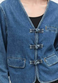 Veste en denim avec un col en V, dotée de fermetures nouées, de deux poches avant et de coutures contrastées. Le tissu est bleu moyen et légèrement délavé.