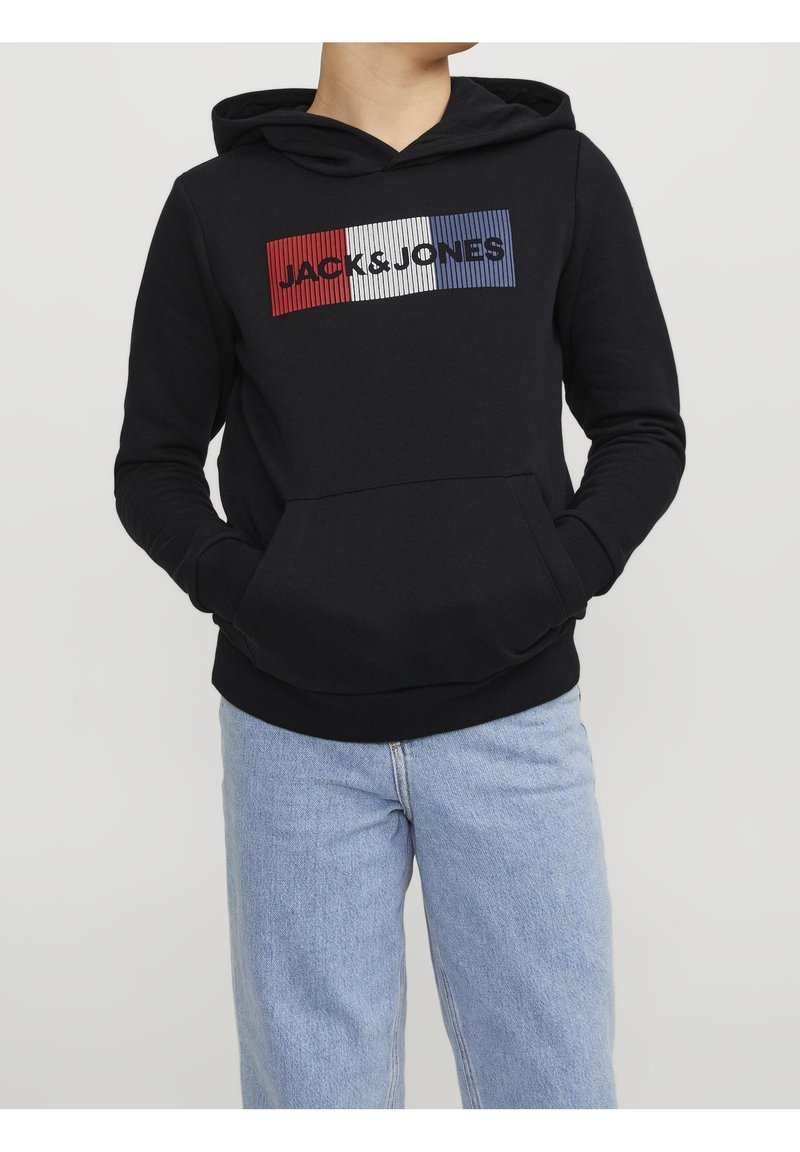 Personne portant un sweat à capuche noir avec le logo rouge, blanc et bleu « JACK & JONES », associé à un jean bleu clair, mains dans les poches du sweat.