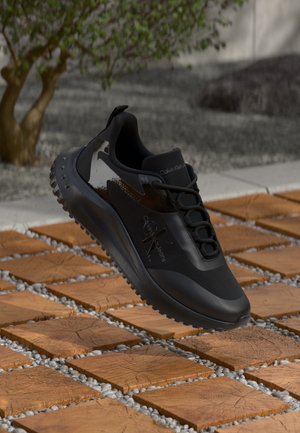 Black athletic shoe featuring a smooth upper with textured accents, a padded collar, and a patterned rubber sole, suspended above a textured surface.