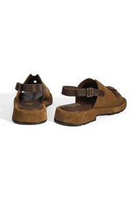 Braune Wildleder-Sandalen mit einer dicken Sohle, offener Ferse, verstellbarem Riemen und einem strukturierten Fußbett. Mit einem dunkelbraunen Akzent auf dem Riemen.