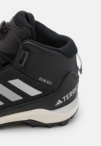 Svart vandringskänga med texturerad ovandel, silverdetaljer och en framträdande logotyp. Har en hög ankeldesign och en robust sula för bra grepp.