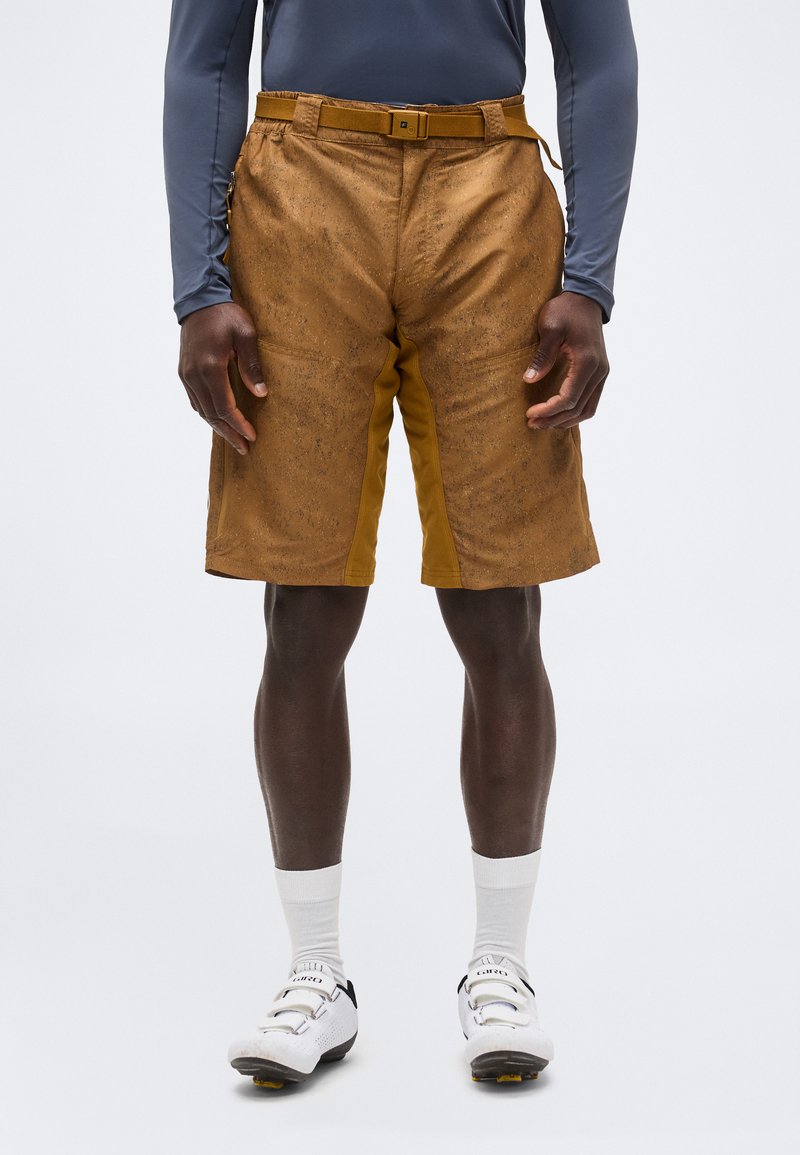 Καφέ ποδηλατικό σορτς με υφή και ενσωματωμένη ζώνη, φορεμένο από ένα άτομο με λευκά ποδηλατικά παπούτσια και λευκές κάλτσες, που στέκεται μπροστά σε απλό φόντο.