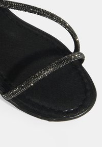 Schwarze Sandale mit einem oberen Teil aus weichem Stoff, versehen mit dünnen Riemen, die mit schwarzen Strasssteinen verziert sind, und einer strukturierten schwarzen Sohle.