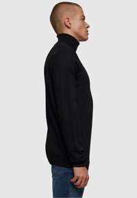 Svart høyhalset genser i slim fit, med lange ermer og ribbestrikket tekstur, parret med blå jeans. Sidesyn.
