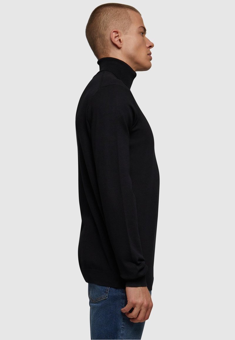 Svart høyhalset genser i slim fit, med lange ermer og ribbestrikket tekstur, parret med blå jeans. Sidesyn.