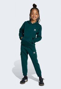 Tumši zaļš kapuci ar kangaroo kabatu un baltu Adidas logo, apvienots ar atbilstošām kargo sporta biksēm. Iezīmes ietver brīvu piegriezumu un ribotas aproces.