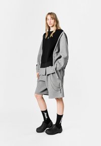 Sudadera gris de gran tamaño sobre una camiseta negra, combinada con pantalones cortos grises y zuecos negros. El atuendo presenta detalles de cadena y calcetas negras.