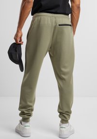 Un pantalon de survêtement vert olive avec une taille élastique, des poches latérales et une poche arrière zippée ; associé à des baskets blanches avec des accents verts.