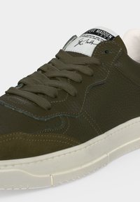 Baskets en cuir vert olive avec surface texturée, lacets assortis, semelle blanche et étiquette blanche sur la languette avec texte noir et signature.
