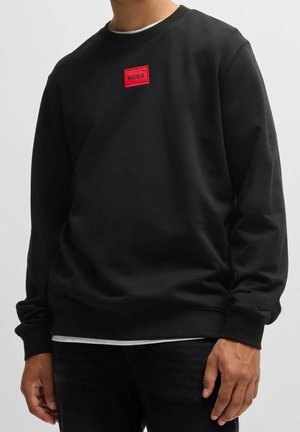 Sweatshirt noir avec col rond, poignets et ourlet côtelés. Présente un patch logo rectangulaire rouge avec du texte en relief sur la poitrine. Matière en mélange de coton.