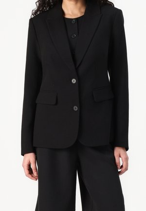 Vrouw draagt een zwarte, op maat gemaakte blazer met twee knopen en klepzakken, gecombineerd met zwarte, wijd uitlopende broek.