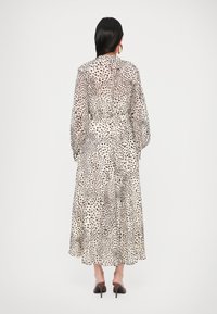 Femme aux longs cheveux foncés portant une robe blanche à pois noirs, manches longues, et des chaussures noires à talons hauts, de dos.