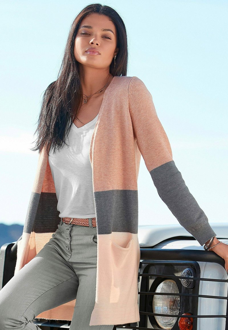 Langer Cardigan in Pfirsich und Grau mit einer leichten Textur. Besitzt einen offenen Vorderteil, zwei Seitentaschen und Streifenmuster. Kombiniert mit einem weißen Oberteil.