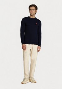 Giovane uomo che indossa un maglione blu navy con logo rosso, pantaloni color crema e sneakers beige, in piedi contro uno sfondo chiaro e uniforme.