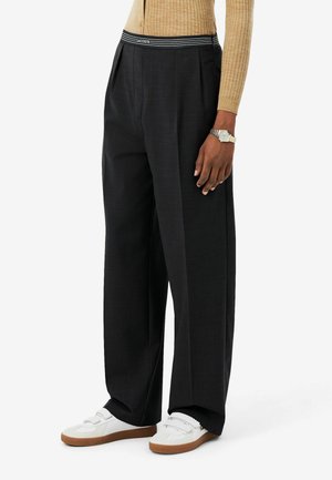 Personne portant un pantalon noir large avec une ceinture rayée Lacoste, un haut beige côtelé à manches longues, des baskets blanches, et une montre argentée.