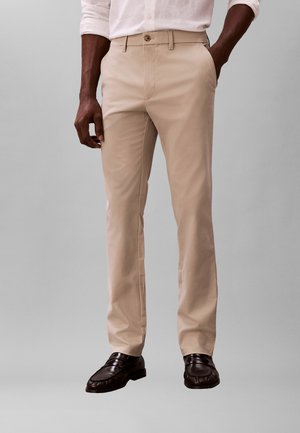 Mann trägt beige maßgeschneiderte Hose mit Seitentaschen, ein weißes Hemd mit Knöpfen und dunkelbraune Loafers, steht vor einem einfarbigen Hintergrund.