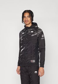 Črna zip-up kapuca z temnimi strukturalnimi vzorci, belimi grafičnimi akcenti in UFC logotipom. Ima kapuco z vrvico in sprednja žepa.
