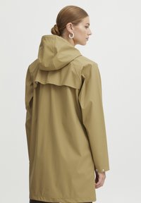 Tan vattentät regnjacka med huva, med en ryggsöm, långa ärmar och tryckknappar vid manchetterna. Slät, matt textur.