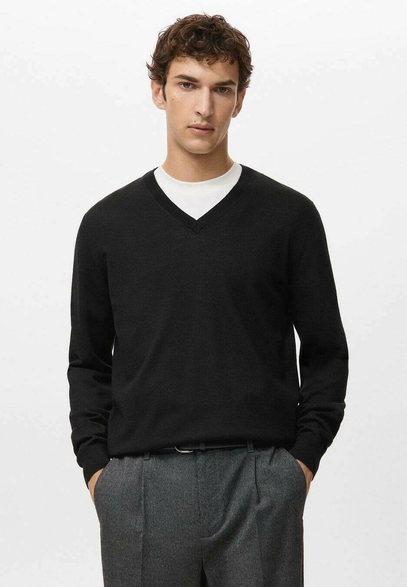 Man draagt een zwarte V-hals trui over een wit overhemd met grijze geplooide broek, staand met handen in de zakken tegen een effen achtergrond.