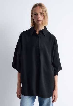 Chemise noire surdimensionnée à boutons avec col, manches courtes et texture douce, portée sur un jean bleu clair.