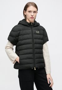 Fekete pufivest rövid ujjakkal és kapucnival, arany logóval a bal mellrészen, cipzáras záródással. Simaszövetű, steppelt design.