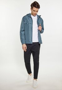 Blå vandafvisende jakke med hætte, frontlynlås og to brystlommer. Kombineret med sorte bukser og hvide sneakers, står foran en lys baggrund.