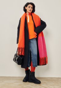 Zwarte wollen jas, oranje trui, spijkerrok met een voorknipt, rode legging, zwarte laarzen en een geleidelijk oranje-roze franje sjaal. Zwarte tas.