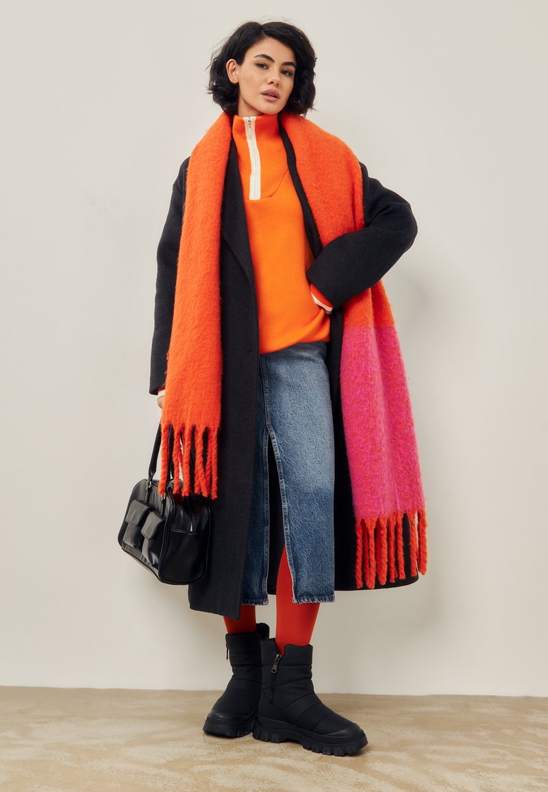 Zwarte wollen jas, oranje trui, spijkerrok met een voorknipt, rode legging, zwarte laarzen en een geleidelijk oranje-roze franje sjaal. Zwarte tas.