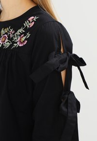 Svart blus med blommig broderi vid halsringningen, öppna axlar med snörningar och en volangkant för extra textur.