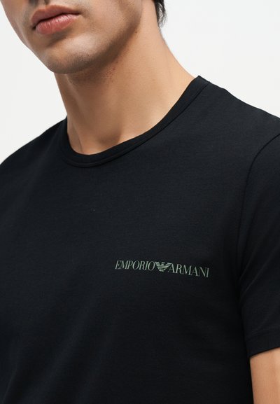 Κοντινό ενός ατόμου που φοράει μαύρο T-shirt με ένα μικρό λογότυπο "Emporio Armani" εκτυπωμένο ανοιχτοπράσινο στο στήθος.