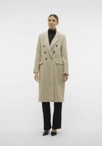 Beige dubbelknäppt kappa med svarta knappar, skräddarsydd design, slagkrage, två framfickor, bärs över en svart turtle-neck och byxor.