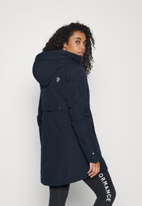 Manteau long bleu marine avec capuche, présentant une fente dans le dos, une fermeture éclair à l'avant, et un logo. Porté sur des leggings noirs avec une écriture blanche.