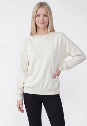 Kobieta z długimi blond włosami, ubrana w kremowy sweter i czarne spodnie, stoi z jedną ręką na biodrze na tle jasnego, jednolitego tła.