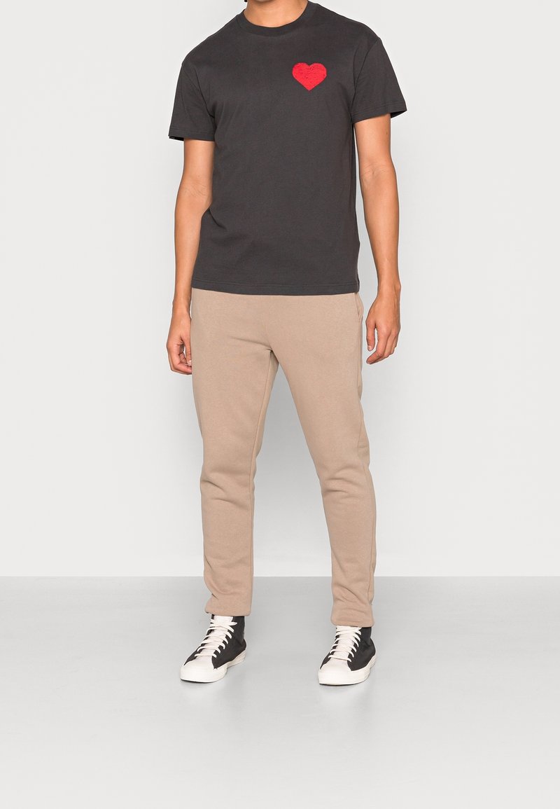 Maglietta nera in cotone con un design a forma di cuore rosso sul lato sinistro del petto, abbinata a pantaloni della tuta beige e sneakers alte in bianco e nero.