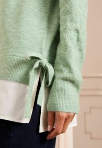 Minzgrüner Pullover mit gerippten Bündchen und geknotetem Saum. Strukturierter Strick mit spitzenähnlichen Mustern an den Ärmeln. Über einem weißen Hemd getragen.