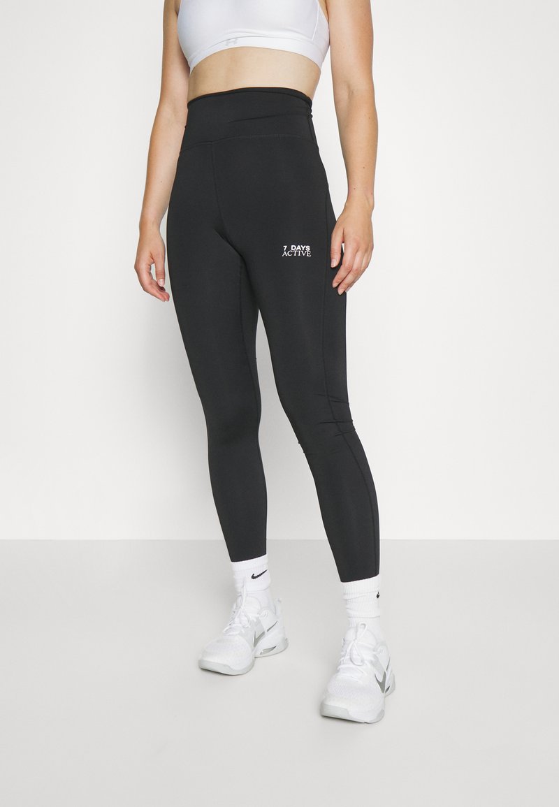 Leggings noirs avec une taille haute, en tissu texturé, portant un logo blanc "7 DAYS ACTIVE" sur le devant, associées à des chaussures de sport blanches.