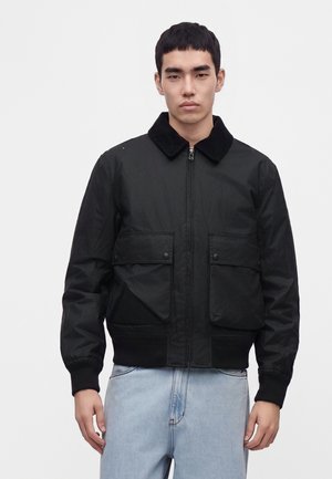 Junger Mann trägt eine schwarze Jacke mit Reißverschluss, Kragen und zwei vorderen Taschen, kombiniert mit hellblauen Jeans, stehend vor einem schlichten Hintergrund.